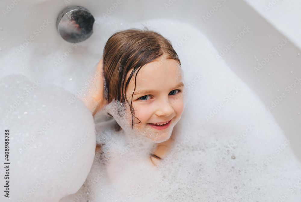 A small, smiling, beautiful red-haired girl with long hair, the child bathes, lying in a white bath with foam from soap and shampoo. Cheerful photography. A small, smiling, beautiful red-haired girl with long hair, the child bathes, lying in a white bath with foam from soap and shampoo. Cheerful photography.