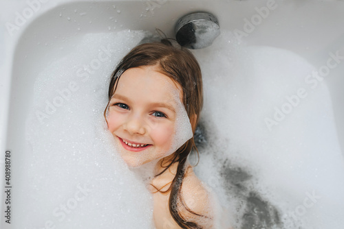 A small, smiling, cute red-haired girl with long hair, the child bathes, lying in a white bath with foam from soap, shampoo and indulges. Model photography.