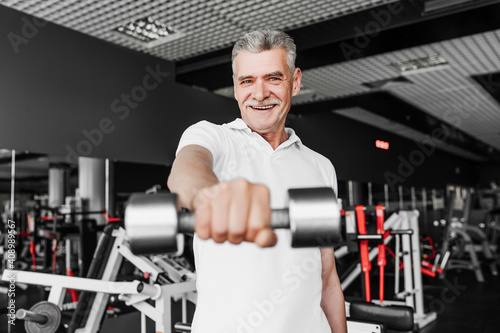 Sport. Close up in focus of a senior man who is smiling and extends his hand with a dumbbell to the camera. Smiling gray-haired man in the gym