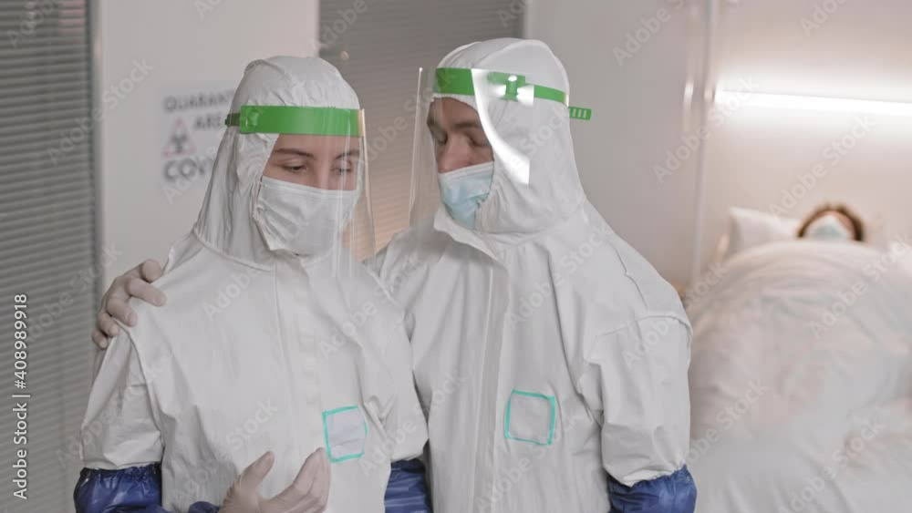 High angle waist-up of doctors wearing white hazmat suits, face shields and masks standing in hospital room. Exhausted woman complaining to colleague, he supporting, holding hand, patting on her head High angle waist-up of doctors wearing white hazmat suits, face shields and masks standing in hospital room. Exhausted woman complaining to colleague, he supporting, holding hand, patting on her head