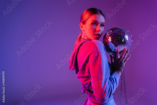 young and stylish woman holding shiny disco ball and looking away on purple