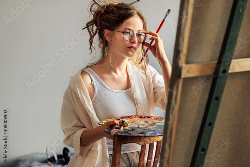 Horizontal candid shot of a girl artist standing next to the easel with canvas painting something in her art studio. A professional young woman painter in transparent eyeglasses draws in the workshop. photo