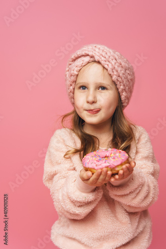 little girl in a fluffy sweater and a headband, smiling with a doughnut in her hand. Sweet appetizer, delicious pastries. a child shows a delicious dessert on a pink background in studio