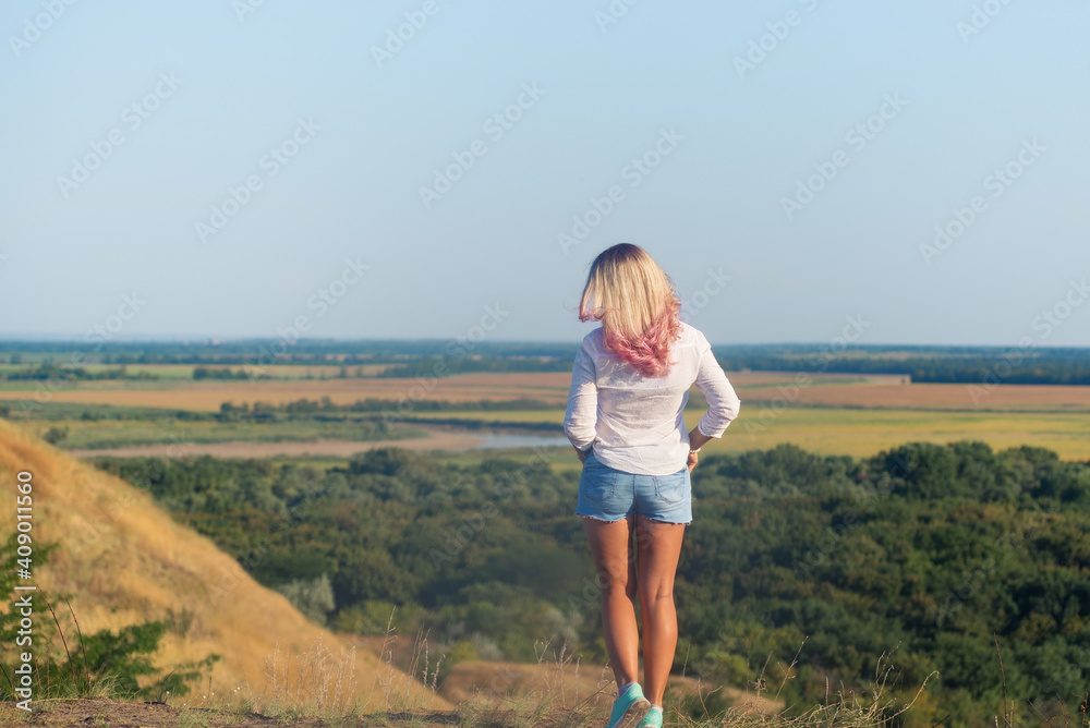 Slim and athletic woman weared white shirt and blue shorts with long blonde pink hair standing on the mountain top over forest and green fields, relax concept