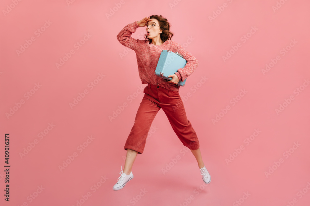 Full length view of brunette woman holding blue valise. Studio shot of caucasian lady with suitcase posing on pink background.