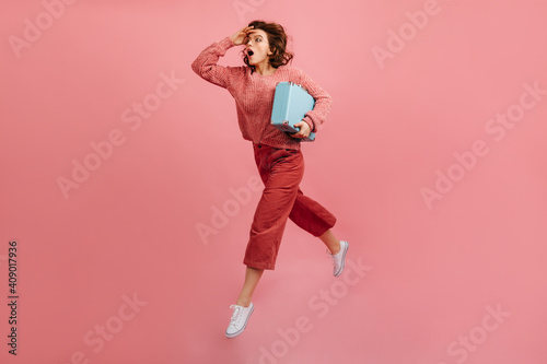 Stressed woman with valise hurrying on pink background. Studio shot of running lady with suitcase.