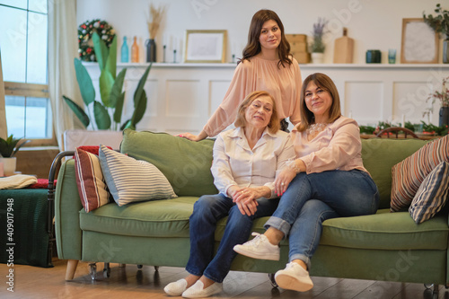 three generations of a family of women are sitting together on the couch. grandmother, daughter, and granddaughter pose together to get a joint photo.