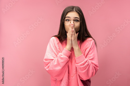 Photo of attractive young woman in glasses is surprised, covers mouth and not expected. Wears casual pink t-shirt white pants isolated pink color background