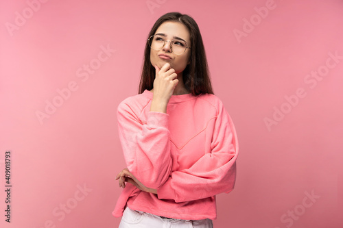 Photo of attractive young woman in glasses thinks and wonder for something. Wears casual pink t-shirt white pants isolated pink color background
