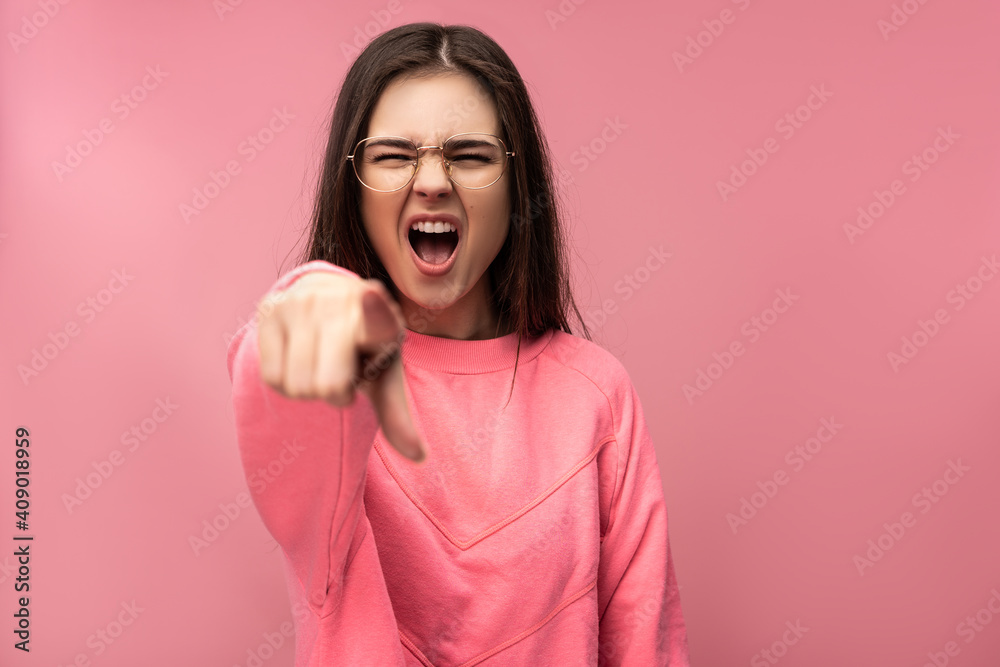 Photo of attractive young woman in glasses yelling and pointing at camera. Wears casual pink t-shirt white pants isolated pink color background Photo of attractive young woman in glasses yelling and pointing at camera. Wears casual pink t-shirt white pants isolated pink color background
