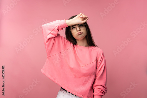 Photo of attractive young woman in glasses looks tired and exhausted, wears casual pink t-shirt white pants isolated pink color background