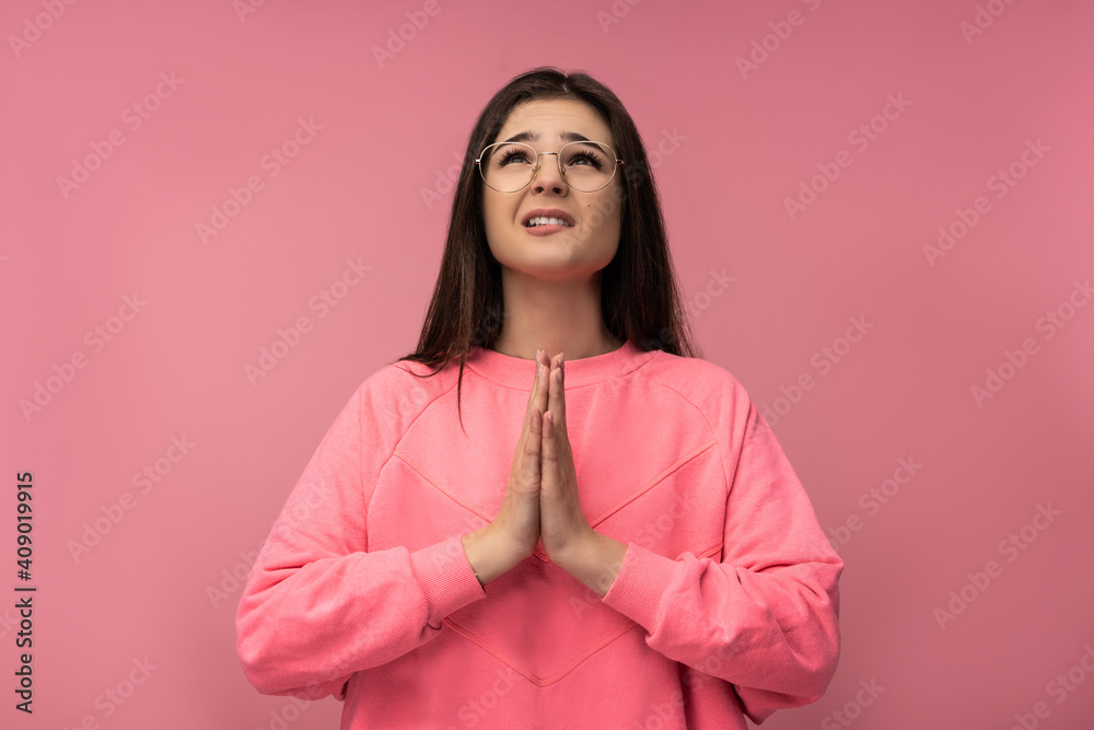 Photo of attractive young woman in glasses uncertainly praying , wears casual pink t-shirt isolated pink color background Photo of attractive young woman in glasses uncertainly praying , wears casual pink t-shirt isolated pink color background