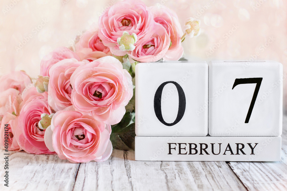 Fototapeta premium White wood calendar blocks with the date February 7th and pink ranunculus flowers over a wooden table. Selective focus with blurred background. 