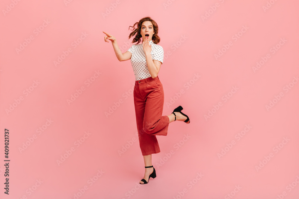 Surprised woman in white t-shirt standing on one leg. Amazed curly lady pointing with finger on pink background.