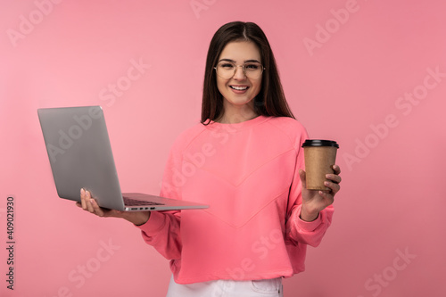 Photo of attractive lady in glasses smile, hold laptop coffee and work online. wear casual pink t-shirt white pants isolated pink color background.