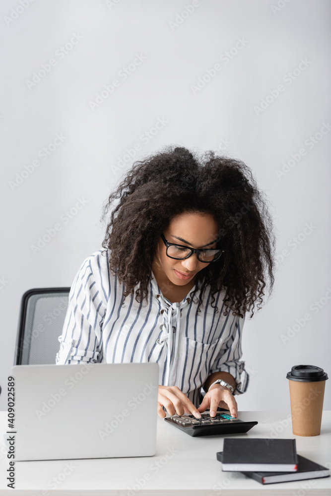 african american freelancer in glasses using calculator while counting near laptop and paper cup on desk - obrazy, fototapety, plakaty 