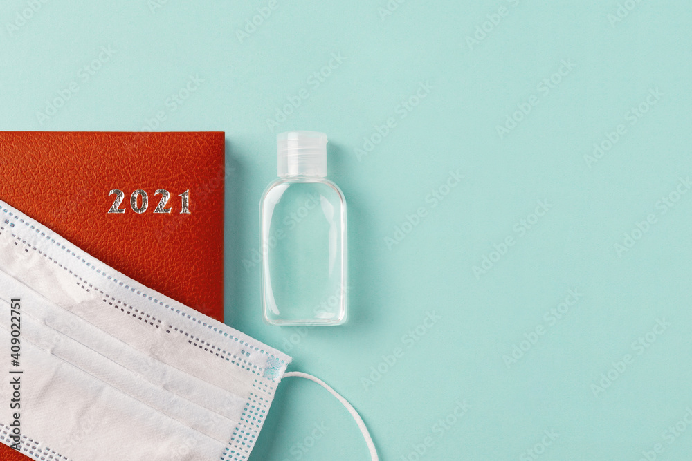 Medical face mask on a 2021 year red leather diary and hand sanitizer on a pale turquoise background. Planning your life and business considering the epidemic. Copy space.