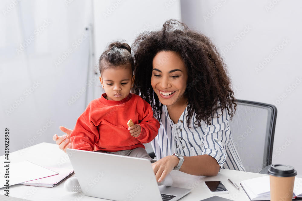 toddler african american child holding tasty cookie and sitting on desk near smiling mother looking at laptop toddler african american child holding tasty cookie and sitting on desk near smiling mother looking at laptop