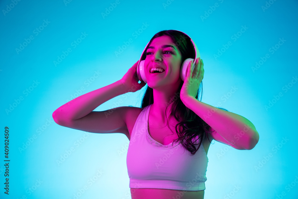 Joyful. Brunette woman's portrait on blue studio background in mixed neon. Beautiful model with headphones listens to music. Concept of human emotions, facial expression, sales, ad, fashion. Joyful. Brunette woman's portrait on blue studio background in mixed neon. Beautiful model with headphones listens to music. Concept of human emotions, facial expression, sales, ad, fashion.