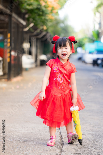 Beautiful child wearing red Chinese clothes takes walk on holiday of traditional Chinese New Year.Baby with sweet smile and bright laugh, stand on street with umbrella.Half Thai-Chinese kid, 3-4 years