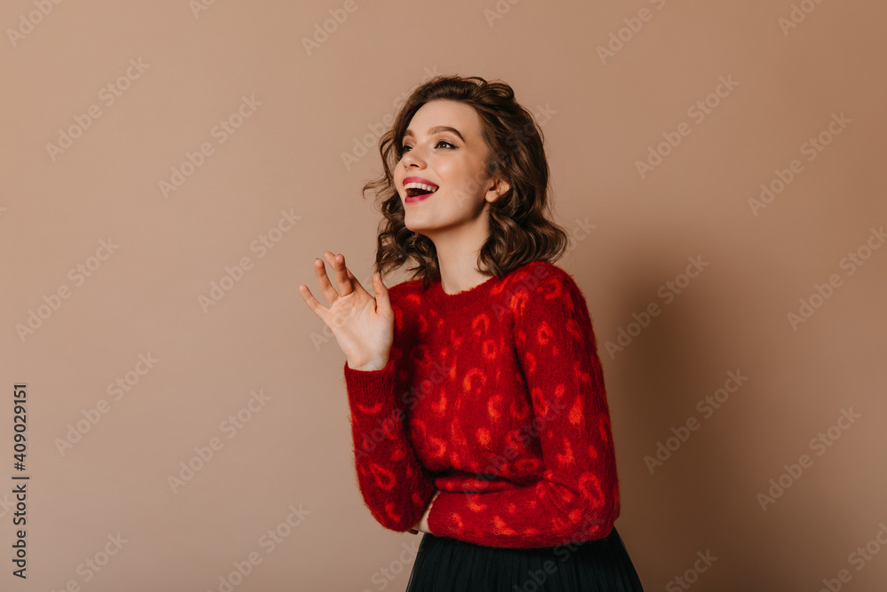 Good-humoured girl with wavy hairstyle smiling on brown background. Studio shot of gorgeous lady in red sweater. Good-humoured girl with wavy hairstyle smiling on brown background. Studio shot of gorgeous lady in red sweater.