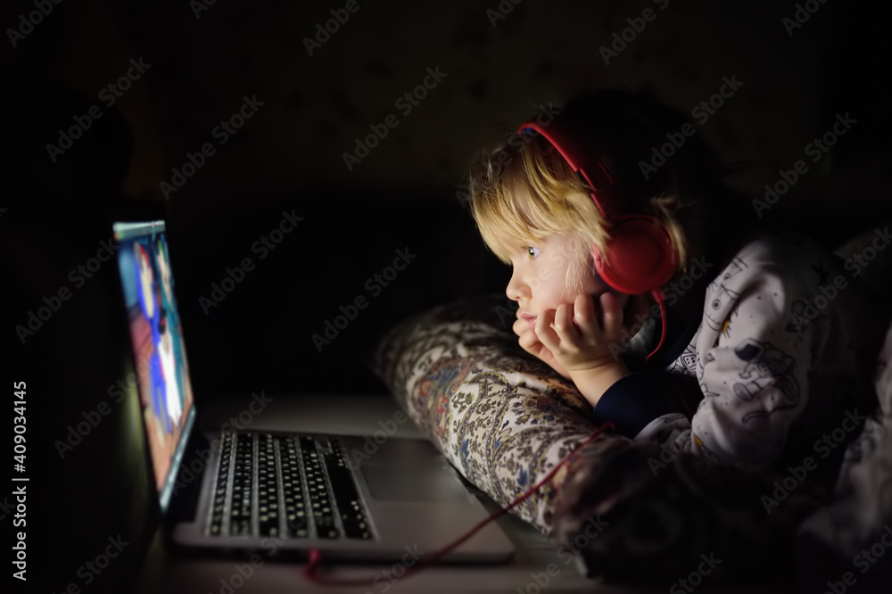 Cute preschool boy wearing headphones is watching cartoon movie by laptop before sleeping in bed or early in the morning right after waking up. Overuse and addiction kids from gadgets. Cute preschool boy wearing headphones is watching cartoon movie by laptop before sleeping in bed or early in the morning right after waking up. Overuse and addiction kids from gadgets.