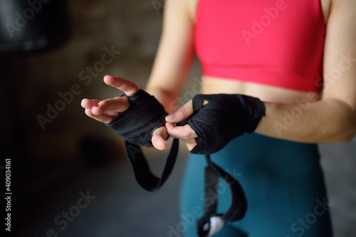 Young female boxer preparing for boxing fight. Athletic woman wearing strap on wrist before boxing practice in gym.