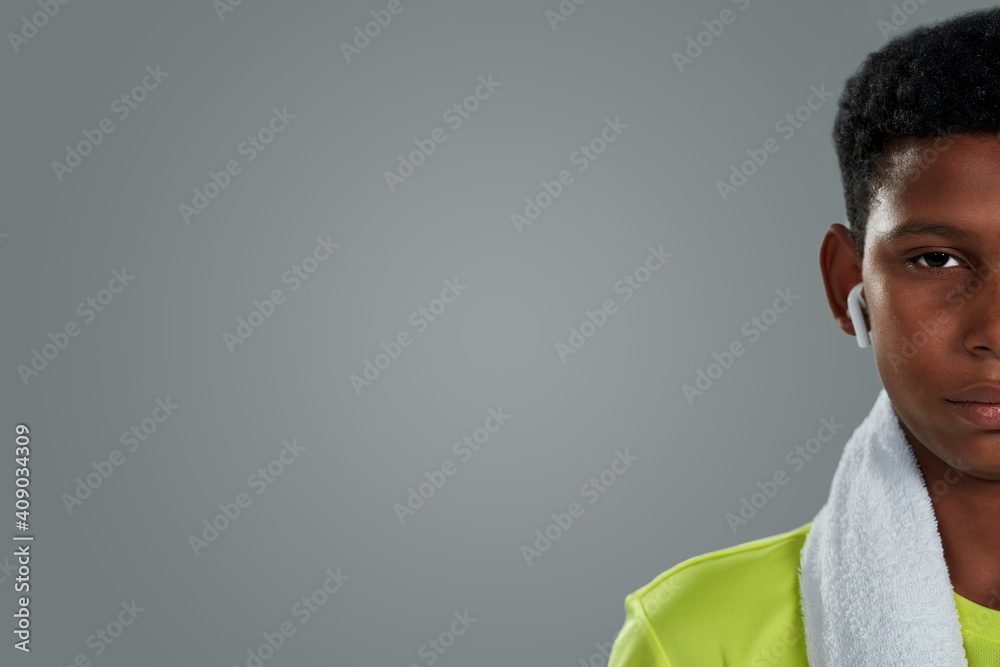 Half face portrait of a confident teenage african boy with white towel on shoulders wearing wireless earphones looking at camera while posing against grey background Half face portrait of a confident teenage african boy with white towel on shoulders wearing wireless earphones looking at camera while posing against grey background