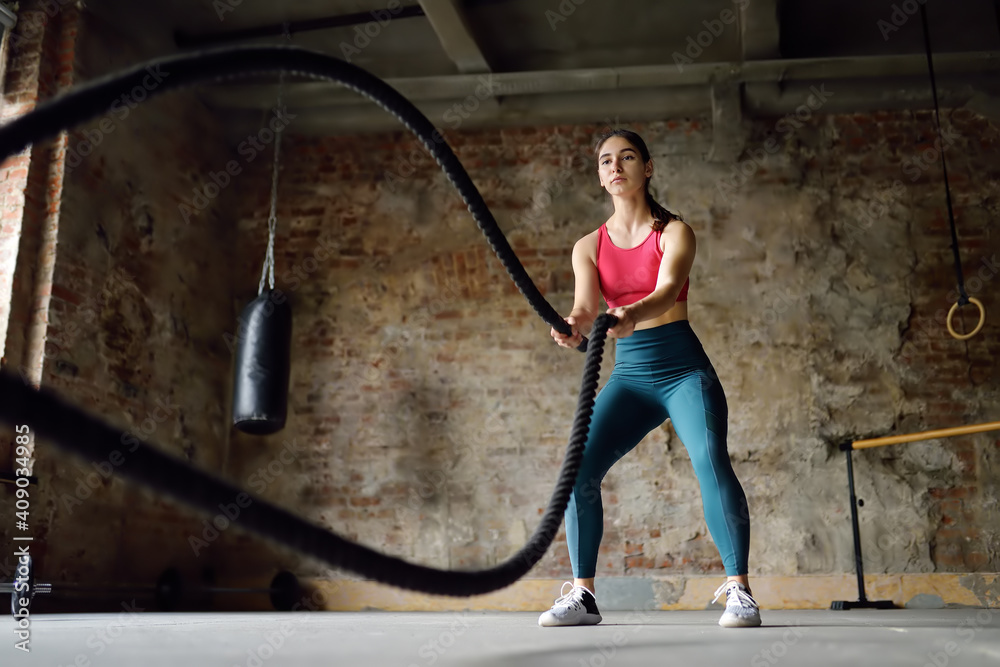 Young woman exercising with battle ropes at the gym. Athletic girl wearing red t-shirt training with battle ropes indoors.