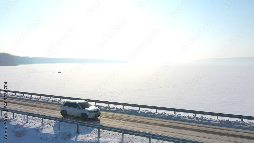 Aerial shot of white car riding through snow covered road near a frozen lake. SUV driving at countryside route near icy pond at sunny winter day. Flying over the auto moving through scenic landscape Aerial shot of white car riding through snow covered road near a frozen lake. SUV driving at countryside route near icy pond at sunny winter day. Flying over the auto moving through scenic landscape