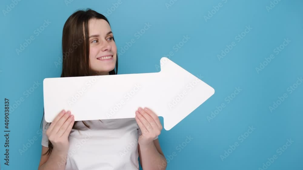 Portrait of emotive young woman hides face behind white paper arrow and points sideways, dressed in t-shirt, isolated on blue background with copy space for advertisement. People emotion concept Portrait of emotive young woman hides face behind white paper arrow and points sideways, dressed in t-shirt, isolated on blue background with copy space for advertisement. People emotion concept