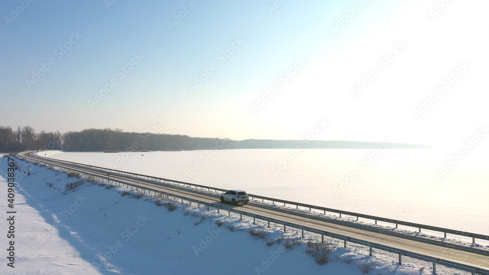 Aerial shot of car riding through snow covered road near frozen lake. White SUV driving at dam route on winter day. Flying over the auto moving through bridge of river. Scenic landscape way. Top view
