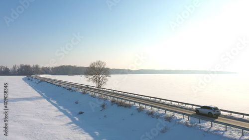 Aerial shot of car riding through snow covered road near frozen lake. White SUV driving at dam route on winter day. Flying over the auto moving through bridge of river. Scenic landscape way. Top view © olehslepchenko