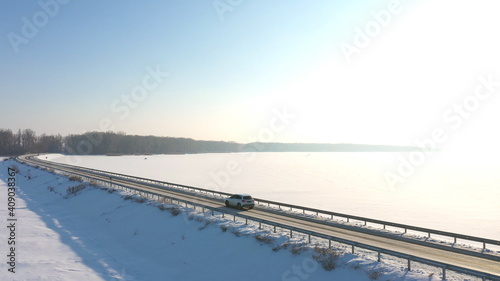 Aerial shot of car riding through snow covered road near frozen lake. White SUV driving at dam route on winter day. Flying over the auto moving through bridge of river. Scenic landscape way. Top view © olehslepchenko