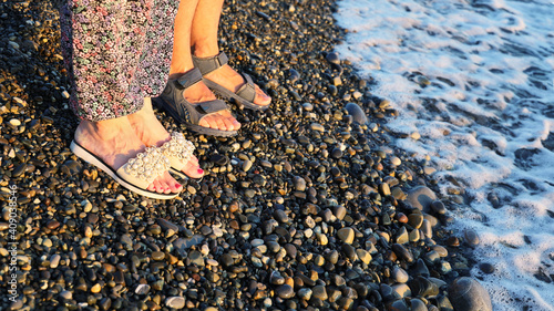 Legs of couple in sandals stand side by side on the pebbled seashore at sunset