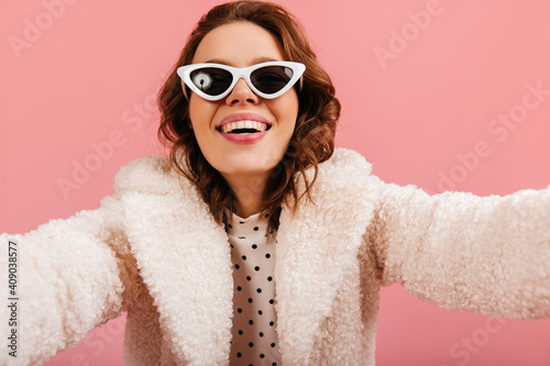 Excited lady in black sunglasses taking selfie. Good-humoured woman in coat isolated on pink background.