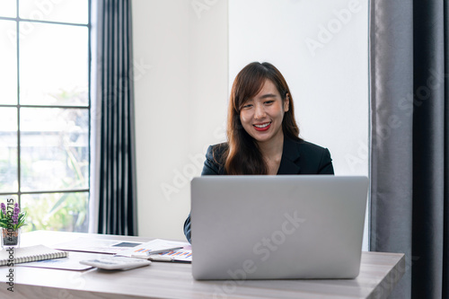 Asian female finance worker working on a laptop At home office table, sit at work with happiness and smile, work from home ideas.