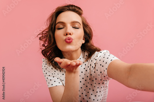 Pleasant girl sending air kiss and taking selfie. Amazing woman expressing love on pink background.