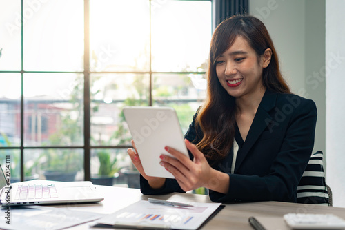 Asian female finance worker sitting at work, answering e-mails on the tablet. At home office table, sit at work with happiness and smile, work from home ideas.