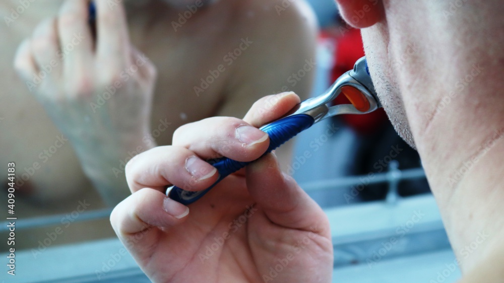 a fragment of the body of a man shaving in the bathroom in front of a mirror close-up, shaving off the stubble from his cheek and chin with a razor, daily male self-care