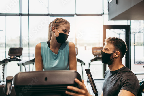 Young fit and attractive woman at body workout in modern gym together with her personal fitness instructor or coach. They keeping distance and wearing protective face masks. Coronavirus sport theme.