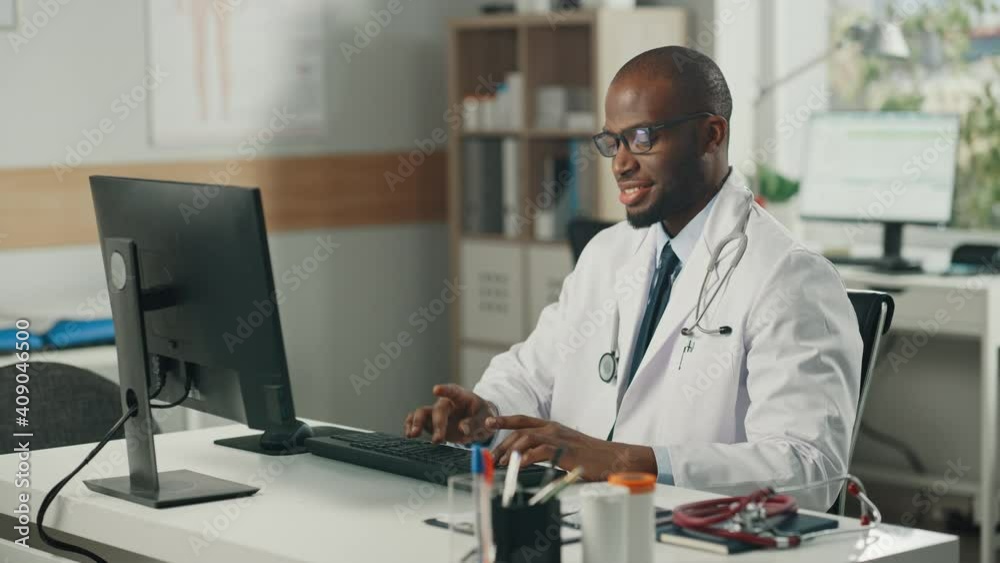 Experienced African American Male Doctor Wearing White Coat Working on Personal Computer at His Office. Medical Health Care Professional Working with Test Results, Patient Treatment Planning. Experienced African American Male Doctor Wearing White Coat Working on Personal Computer at His Office. Medical Health Care Professional Working with Test Results, Patient Treatment Planning.