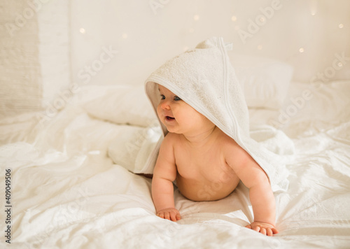 little baby girl in a white towel with a hood is on all fours on a white blanket on the bed