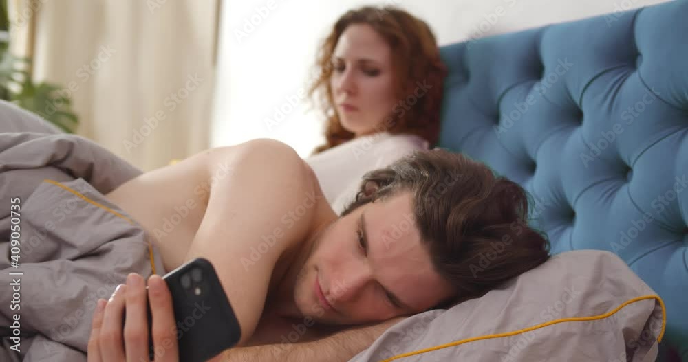 Young couple lying in bed and man cheating on woman chatting on smartphone