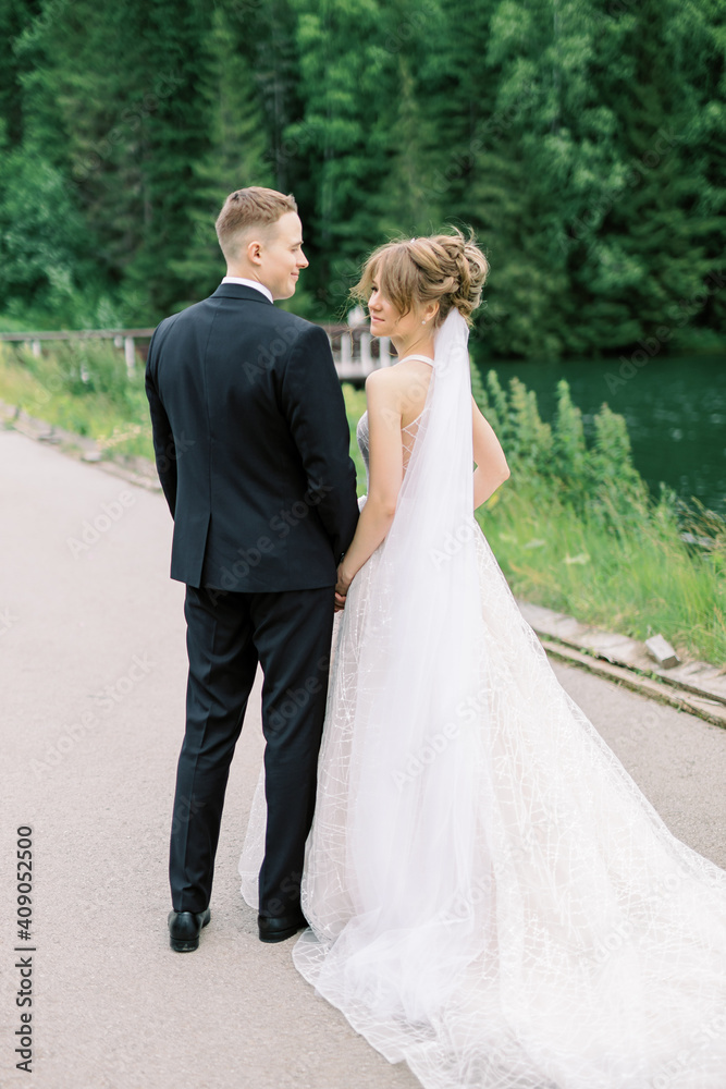 Smiling young woman in wedding dress posing on camera with handsome husband standing behind and holding hands on her waist.