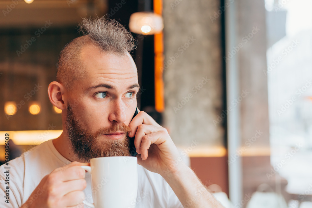 A brutal man of European appearance in a cafe in the morning with a cup of hot coffee speaks by phone A brutal man of European appearance in a cafe in the morning with a cup of hot coffee speaks by phone
