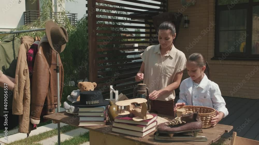  Tracking shot of cheerful woman and girl having garage sale in their front yard on summer day. Bearded man looking at second hand jackets hanging on clothes rack
