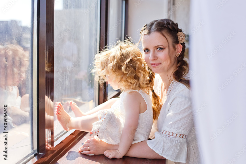 Mother and daughter on the windowsill looking out the window. Happy family at home on the background of a large window. Mother and daughter on the windowsill looking out the window. Happy family at home on the background of a large window.