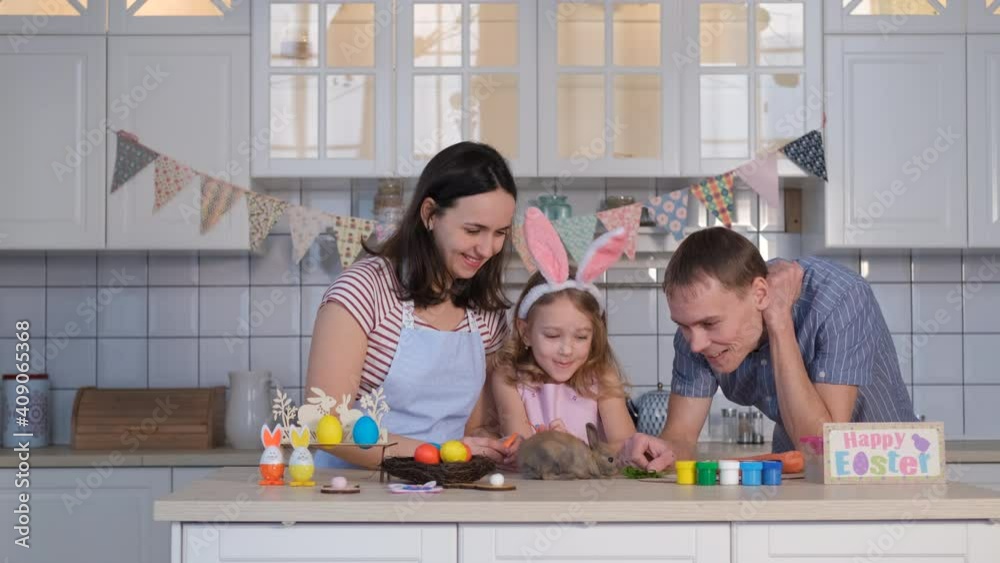 Man with his Wife and Little Daughter Feeding Cute Little Easter Bunny with Greens in Decorated Kitchen. Slow Motion. Family, Easter, Spring, Happiness and People Concept Man with his Wife and Little Daughter Feeding Cute Little Easter Bunny with Greens in Decorated Kitchen. Slow Motion. Family, Easter, Spring, Happiness and People Concept