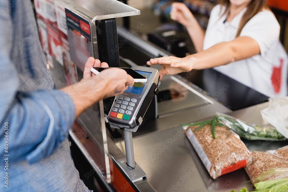 Cropped view of man paying with smartphone near groceries and cashier on blurred background in supermarket Cropped view of man paying with smartphone near groceries and cashier on blurred background in supermarket